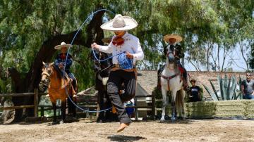 A trick roper in rancho period garb jumps through a rope in front of two horsemen. 