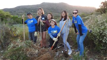 Six students with gardening tools stand surrounded by native plants with a pile of removed weeds in front of them.