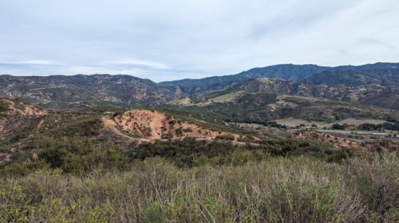 A wide landscape view of rolling hills and rugged mountains covered in green brush and exposed red earth beneath a soft, overcast sky.