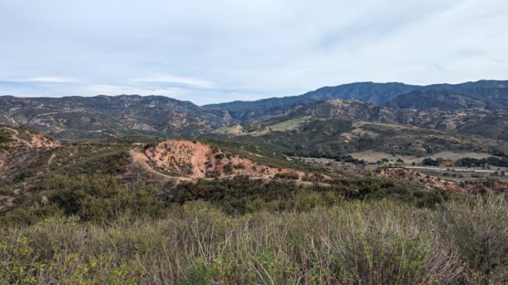 A wide landscape view of rolling hills and rugged mountains covered in green brush and exposed red earth beneath a soft, overcast sky.