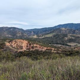 A wide landscape view of rolling hills and rugged mountains covered in green brush and exposed red earth beneath a soft, overcast sky.