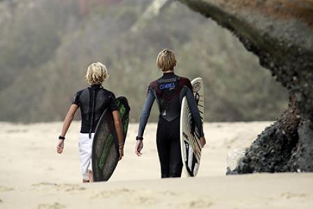 Two young boys stroll on a beach holding surfboards